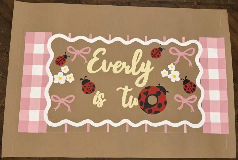 Hand-painted second birthday banner with a pink gingham border, ladybugs, daisies and bows reading Everly is two