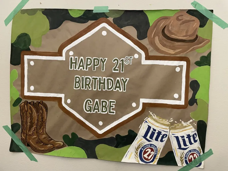 Hand-painted 21st birthday banner reading Happy 21st Birthday Gabe with a camo border, cowboy hat, boots and clinking cans