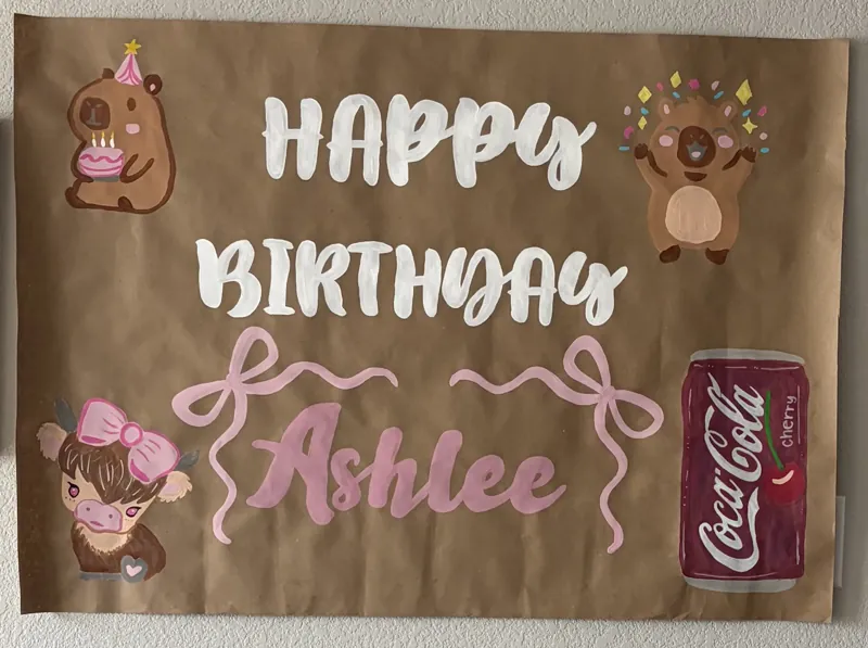 Hand-painted birthday banner reading Happy Birthday Ashlee in pink script with a highland cow, a capybara in a party hat, pink bows and a cola can