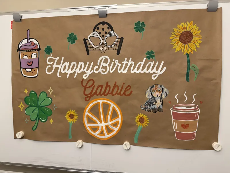 Hand-painted birthday banner on kraft paper reading Happy Birthday Gabbie with sunflowers, a basketball, iced coffee, shamrocks and a dachshund